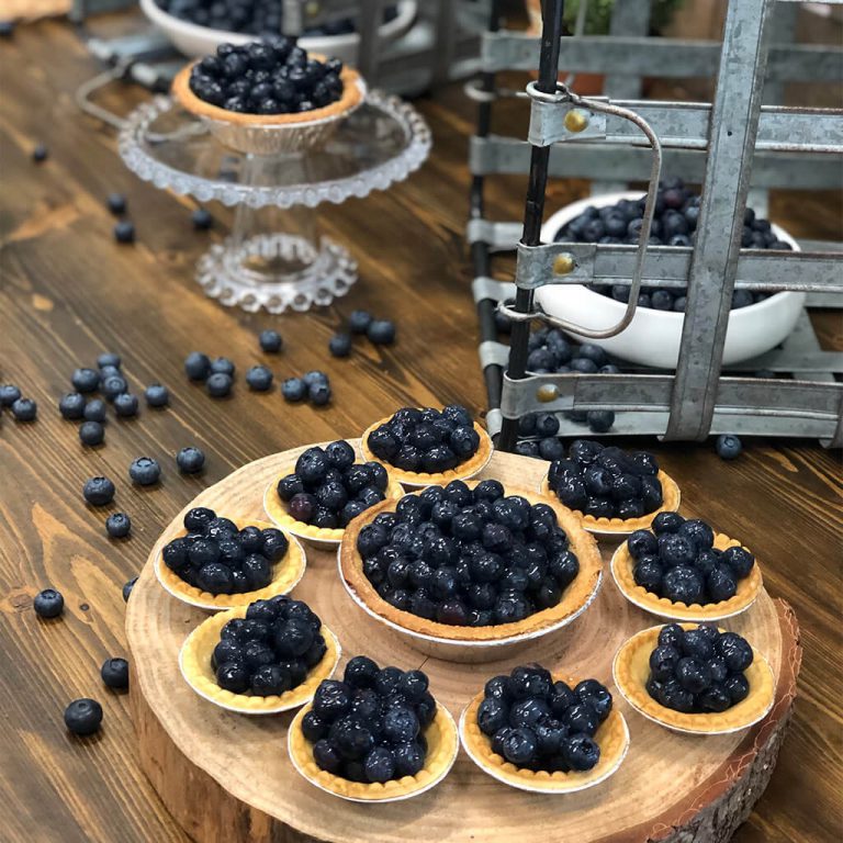 Blueberry tarts arranged on a wood cut charger, with baking supplies and loose blueberries on a wooden table in background.