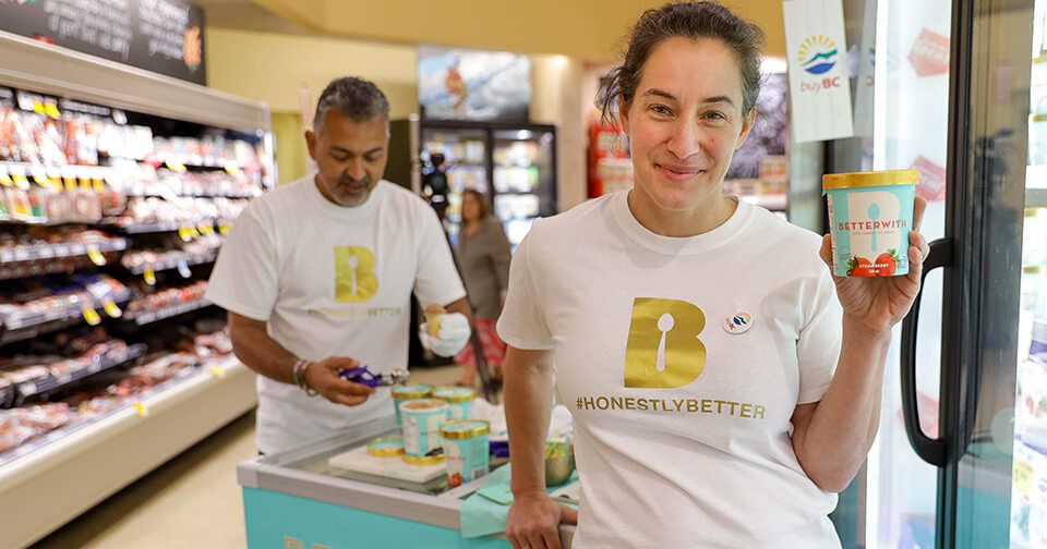 Two people in grocery aisle wearing hashtag Honestly Better tshirts. Person in front holds Better With ice cream container and smiles.