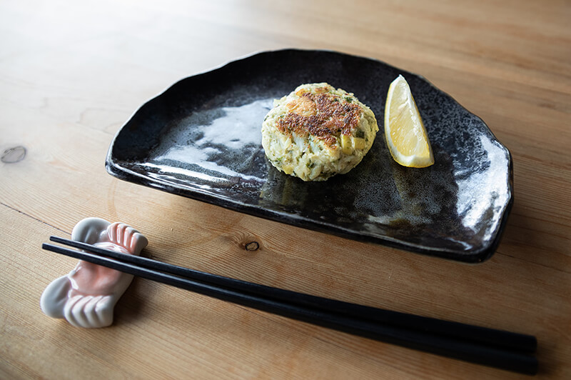 One crab cake next to lemon slice on a semi-circle black dish. Chopsticks rest on a ceramic crab next to the dish.