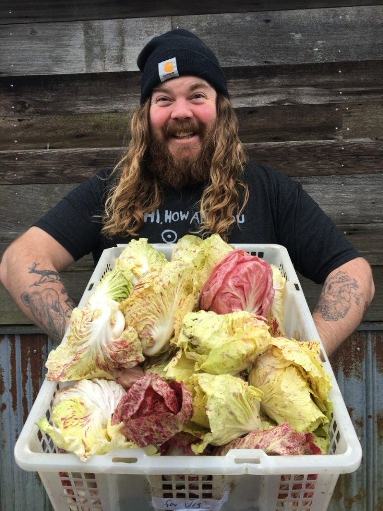 Man with long hair, beard, tattoos, holds large basket of fresh cabbage and smiles at camera.