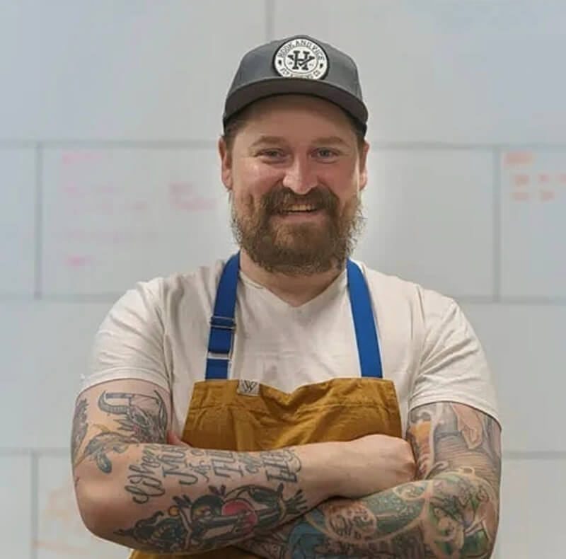 Chef Derek Gray wearing a grey baseball cap and brown and blue apron, with two full sleeve tattoos, smiling at the camera.