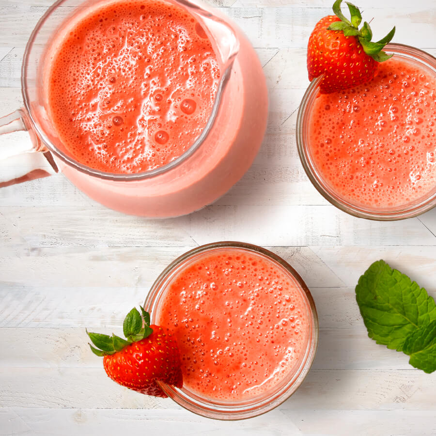 Aerial view of pink strawberry smoothie in two glasses with a strawberry on each glass rim and a pitcher.