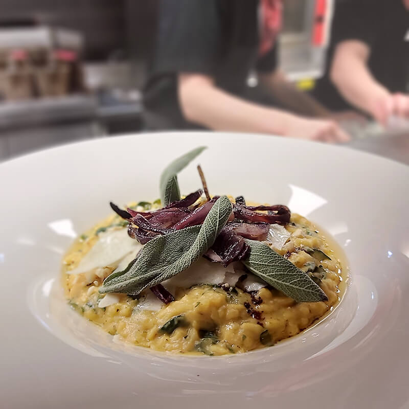 Butternut squash risotto with dried leaves on top in the centre of a wide-rimmed white plate.