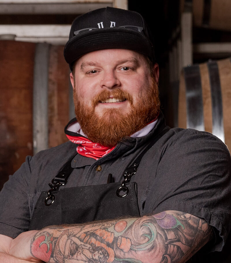 Chef AK in a black hat and apron with full sleeve tattoo, smiling at the camera.