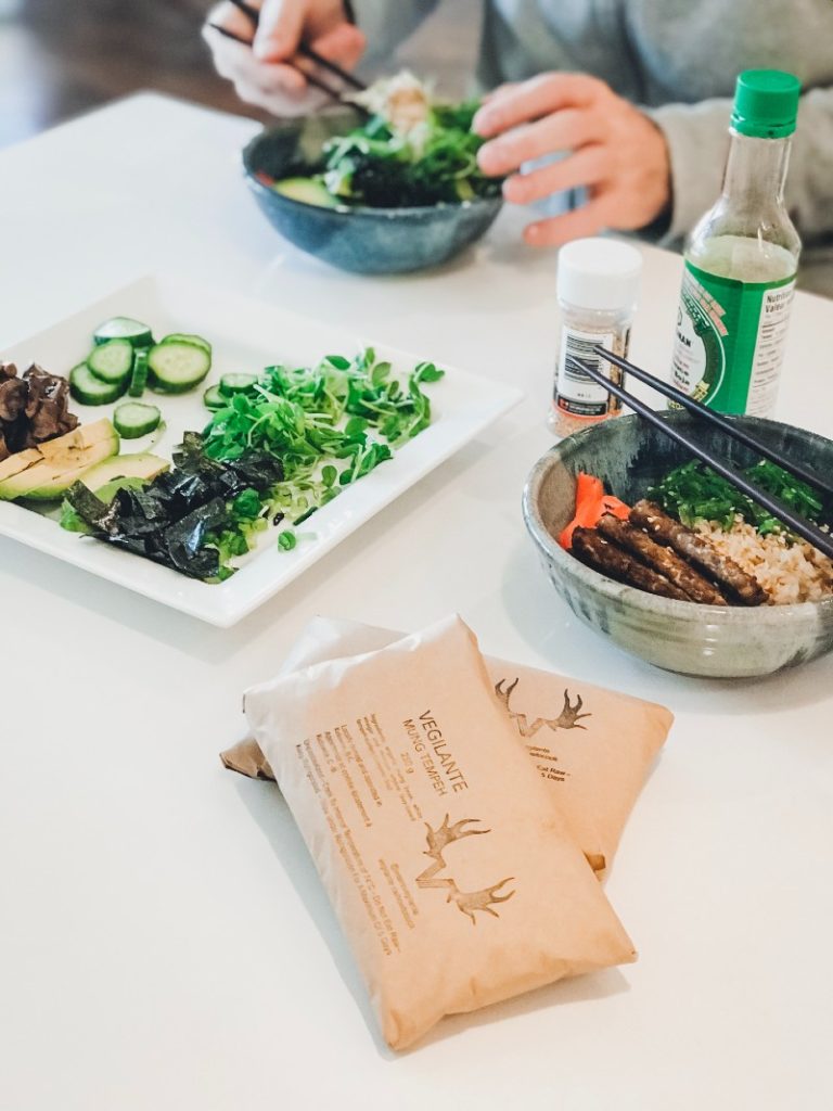 Two packages of vegilante next to someone sitting to eat plated meal of greens, tempeh, mushrooms, and carrots.