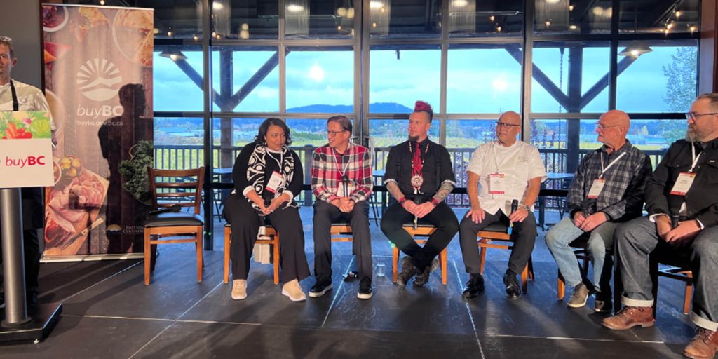 Six panelists sitting on chairs next to a presenter at a podium with Buy BC banners surrounding. Panelists sit in front of windows with farm fields outside.