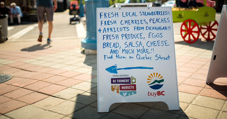 Sandwich board at farmer's market with handwriting, BC Farmers' Markets logo, and Buy BC logo.