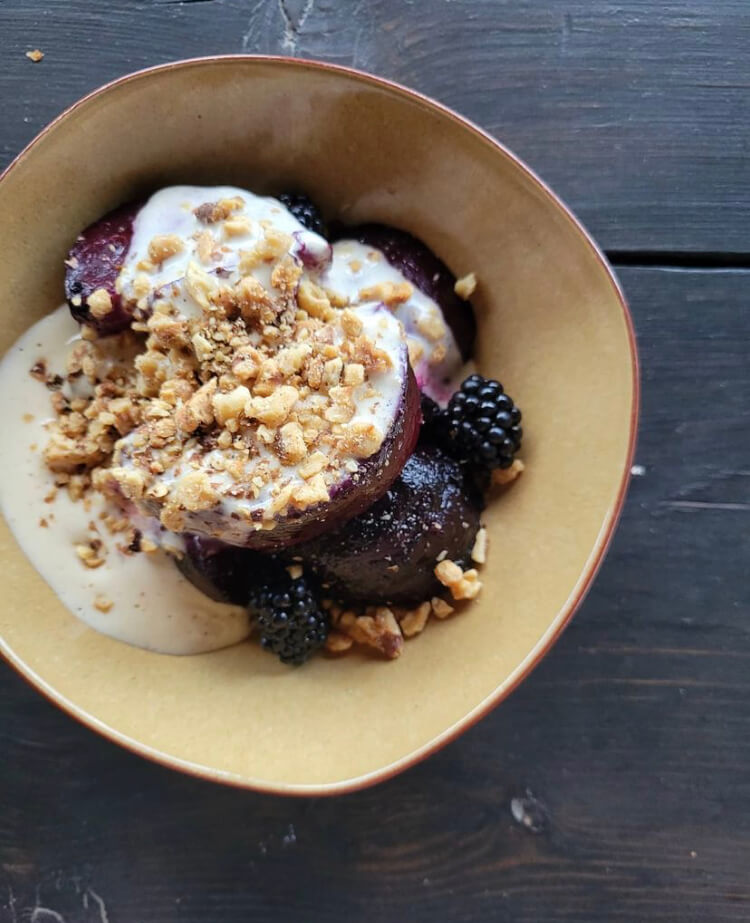 Coal roasted beets by Chef Derek Gray served with blackberries, creamy sauce, and chopped nuts in a light yellow bowl on a dark wood surface.