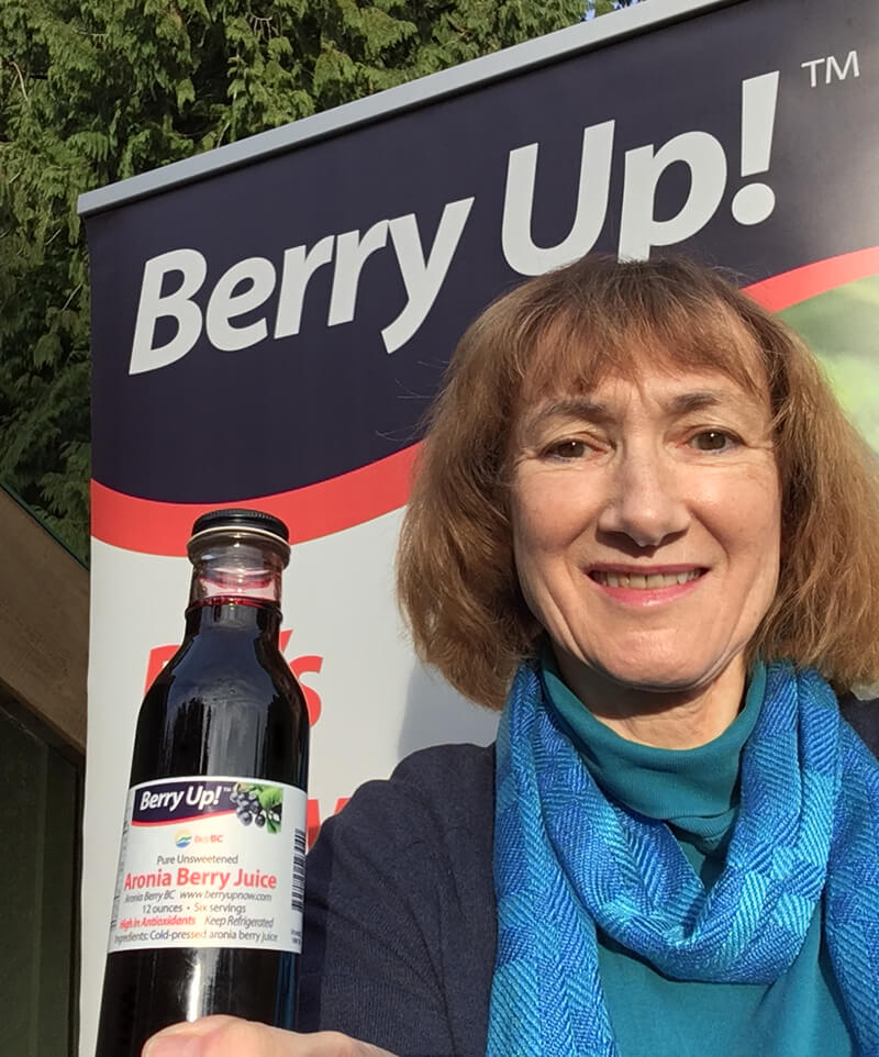 Woman with blue scarf smiles while holding a bottle of berry juice in front of a sign that reads "Berry Up!"