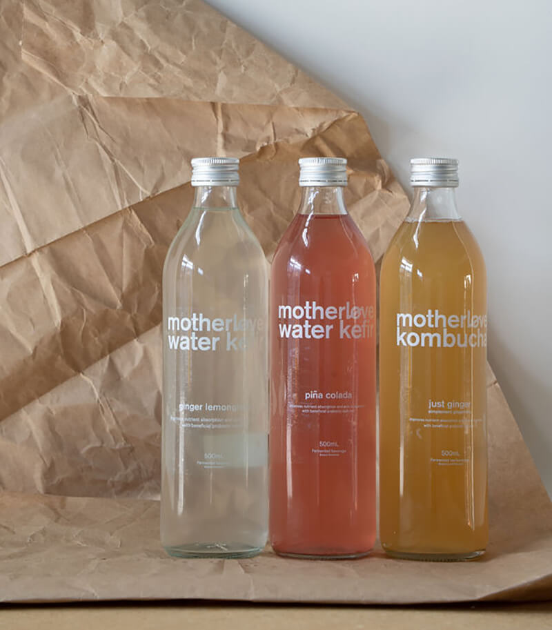 Three glass bottles of kombucha: clear, pink, and yellow, standing on crinkled packaging paper.