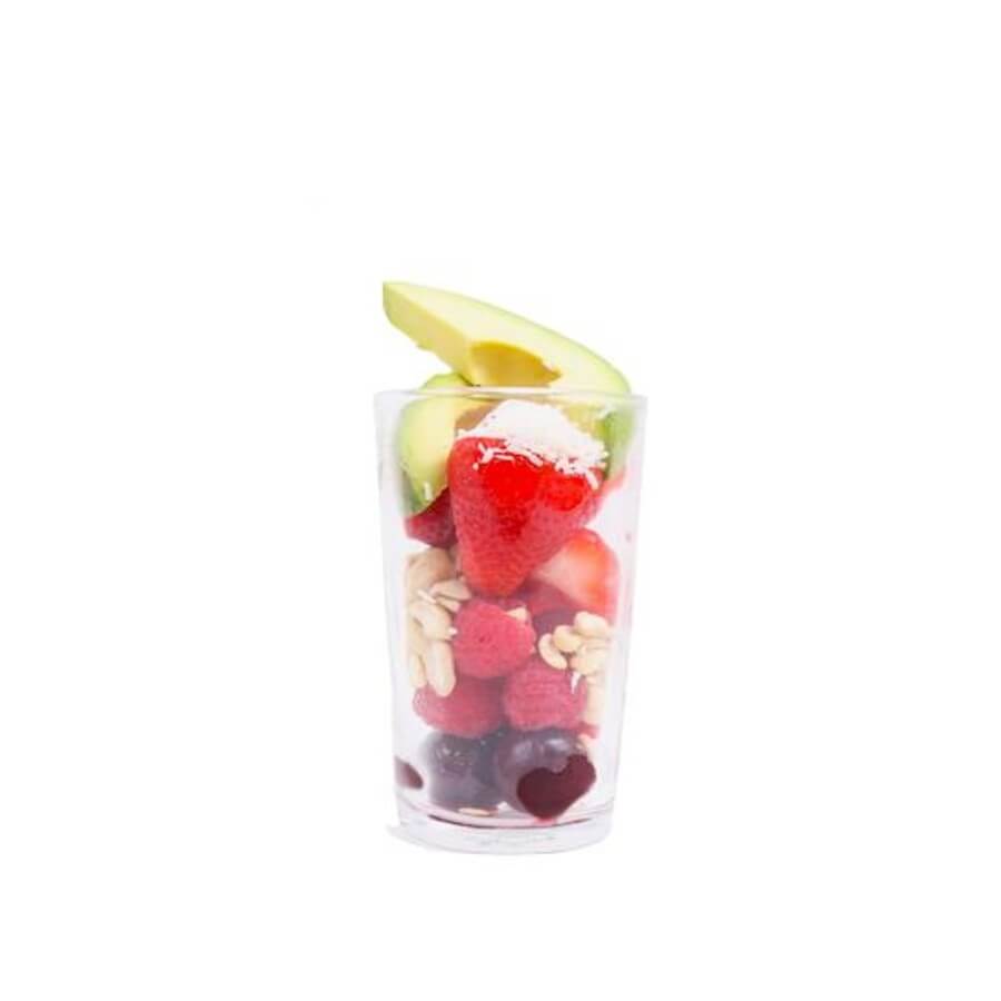 A glass on white background with layered ingredients: Cherries, raspberries, cashews, strawberries, shredded coconut, and avocado.