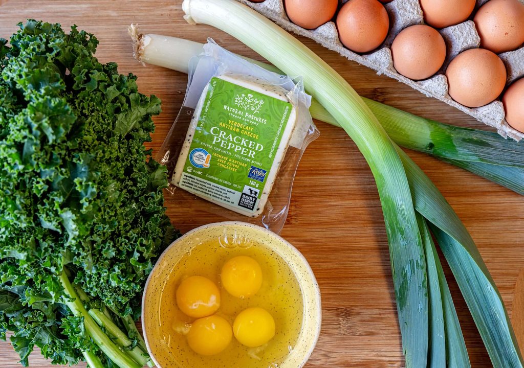 Frittata ingredients: egg yolks and whites in a bowl next to a bunch of B.C. kale and leeks and cracked pepper cheese in the package.