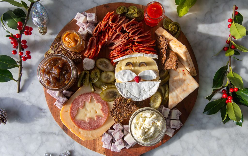 A Holiday cheeseboard with Brie and salami arranged to look like a Santa face and hat with cheese and condiments spread all around it. 