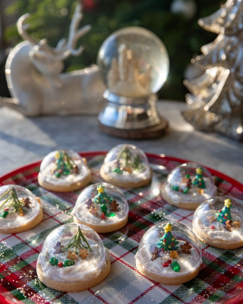 Snow Globe sugar cookies set on a red, green and white platter with a real snow globe in the background.