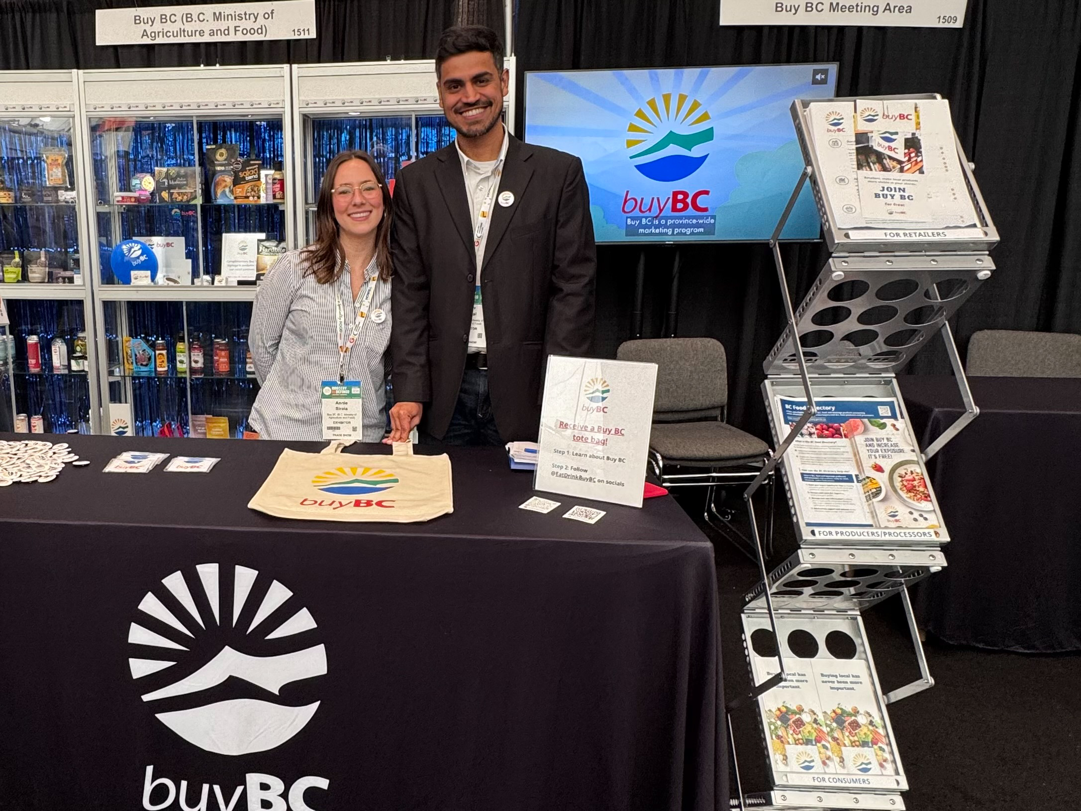 Trade Show Floor Two people standing behind a 6ft table with a black table cloth with the Buy BC logo.