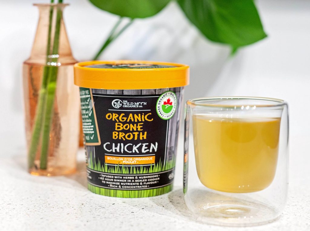 A container of previously frozen The Wild Moon Organics Organic Chicken Bone Broth beside a clear cup of the warm broth on a white background with green plant fronds in a clear orange vase.