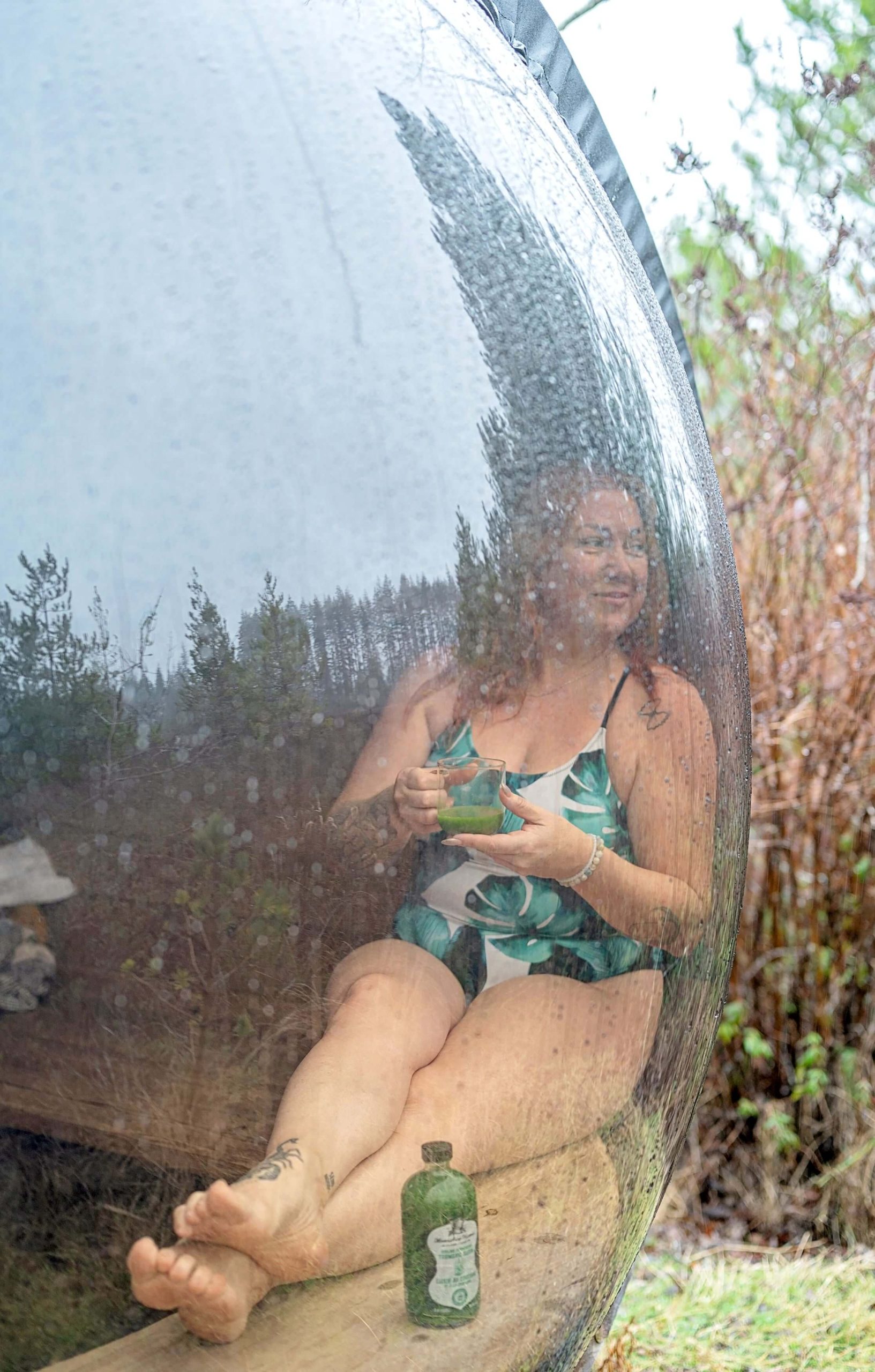 A woman practicing self-care, sitting in a sauna with her feet up on a curved wooden bench, looking out a bubble shaped clear window-wall while sipping a green juice.