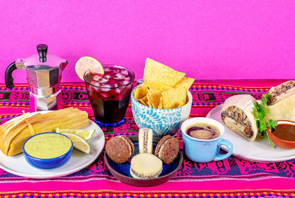 An array of Latin American foods plated and displayed on a vibrant pink blue and yellow Mexican fabric. Supporting Local Latin businesses, all the foods are made or sourced in B.C.