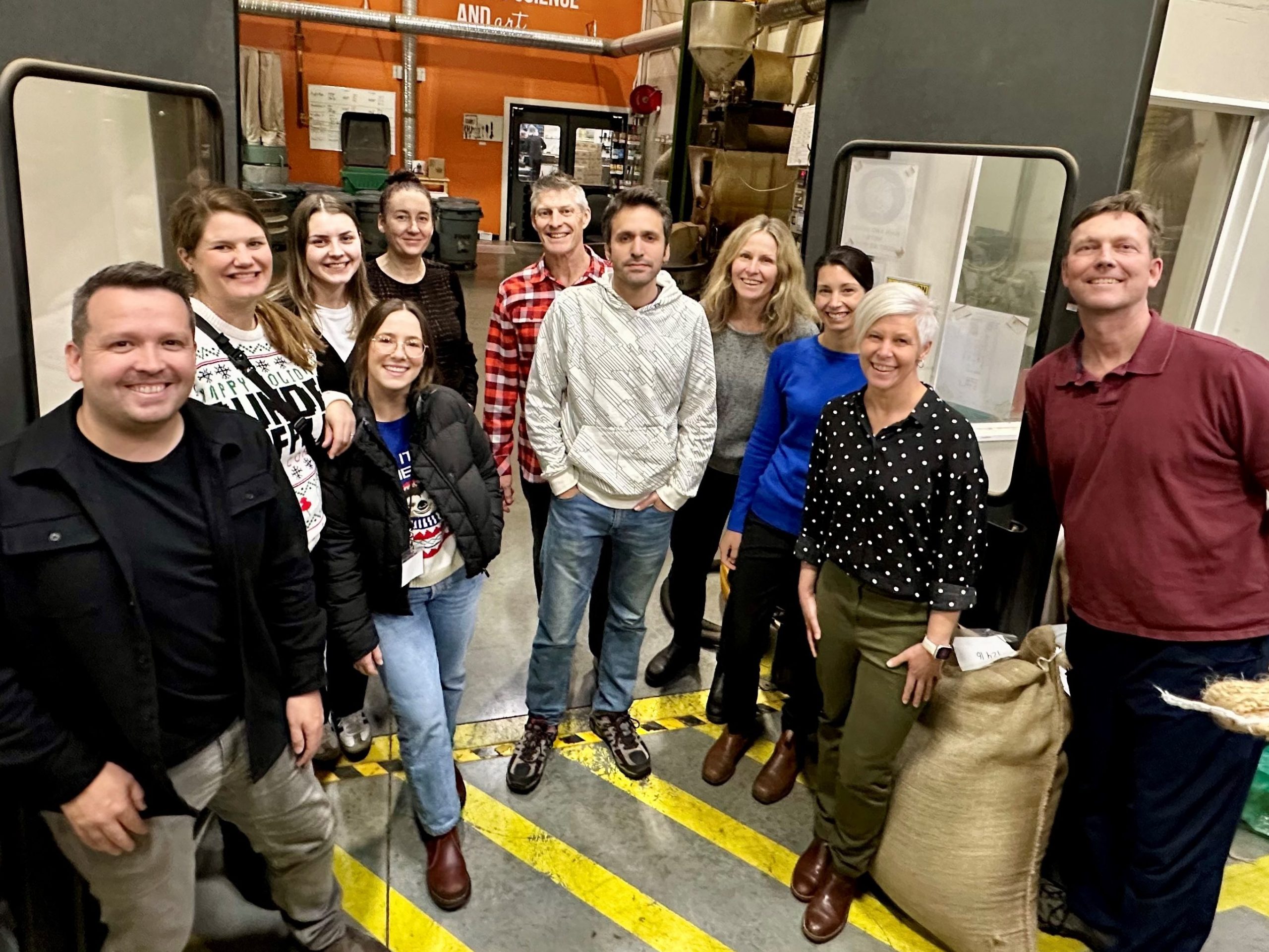 A group of smiling people standing in the door way in the storage area of a B.C. coffee roastery. The first stop on the group's visit to two small local businesses. 