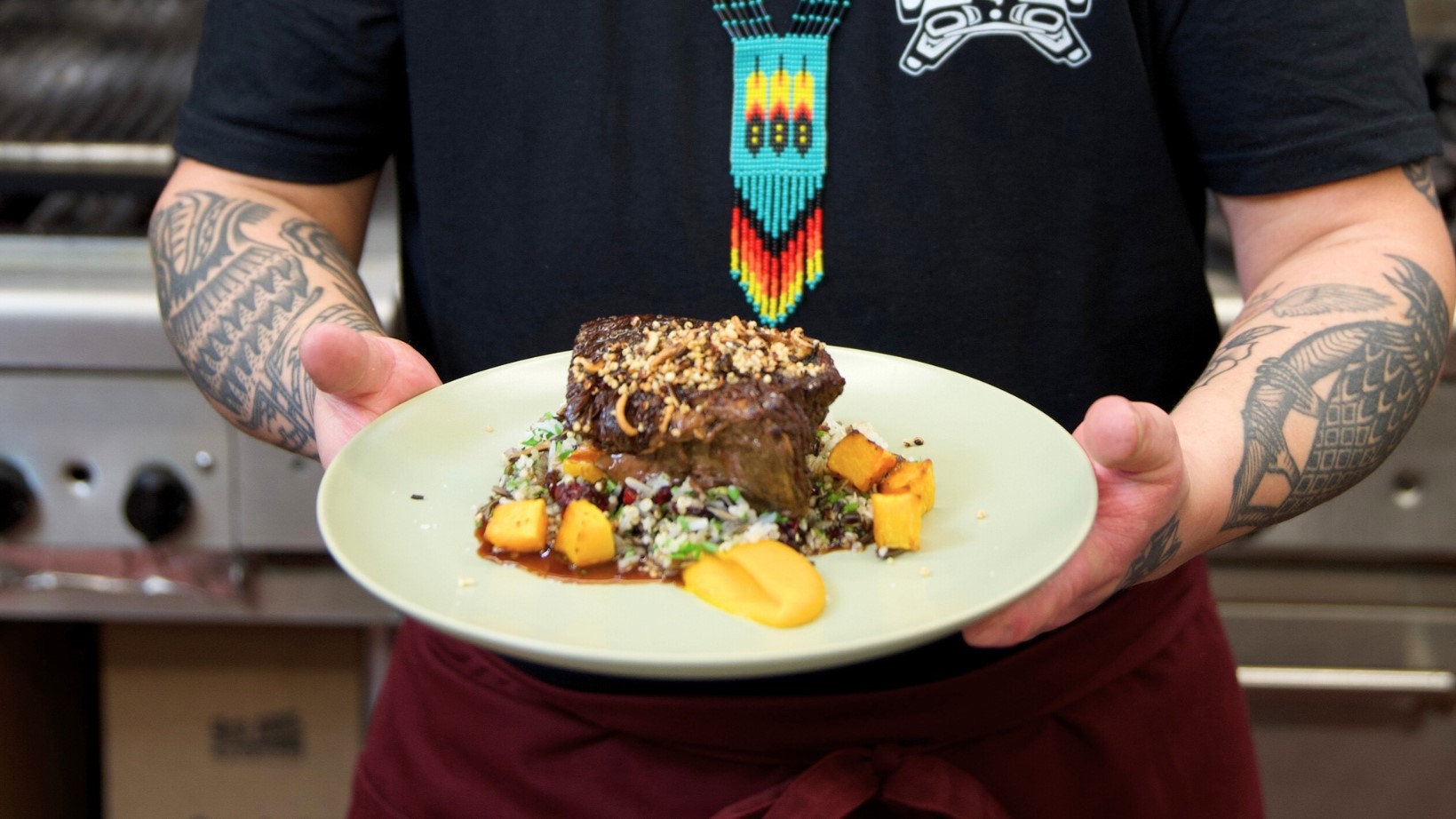 Close up of person with tattooed arms holding a plate with braised bison chuck roll and butternut squash, with an indigenous beadwork neckless in the background on the person's chest.