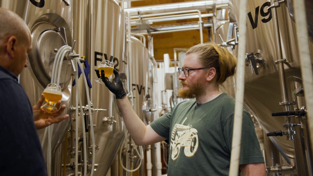 A person holds a glass of B.C. beer near tanks inside a brewery.