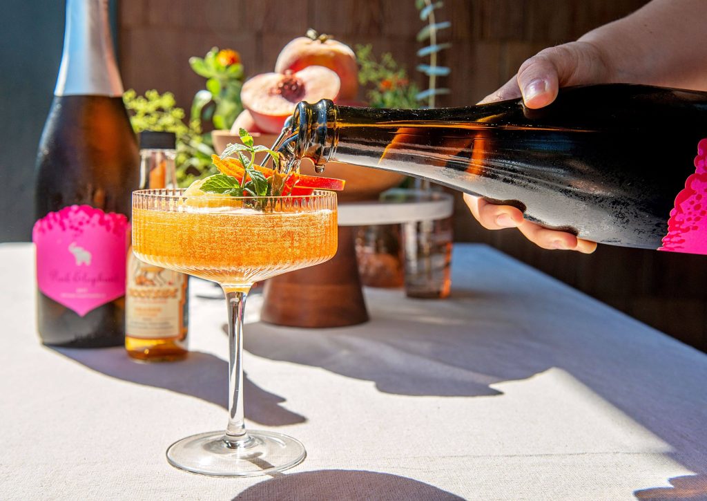 A hand is holding a bottle and pouring sparkling wine into a frozen B.C. peach mixture in a coupe glass sits on a white table cloth. 