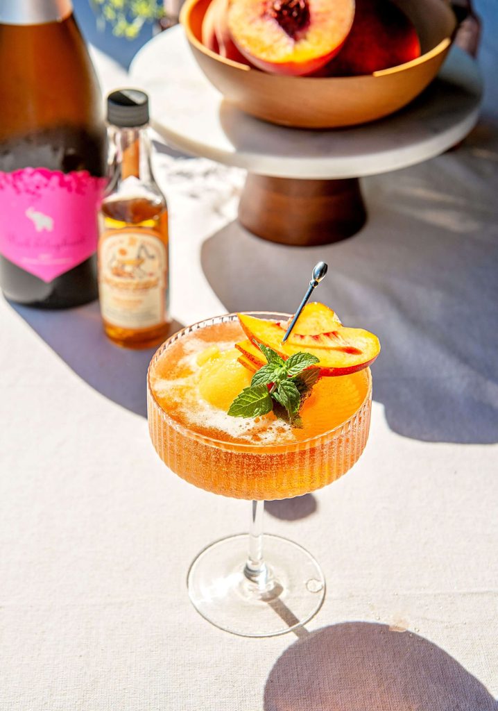 A coupe glass filled with a peach cocktail sits on a white tablecloth outside.