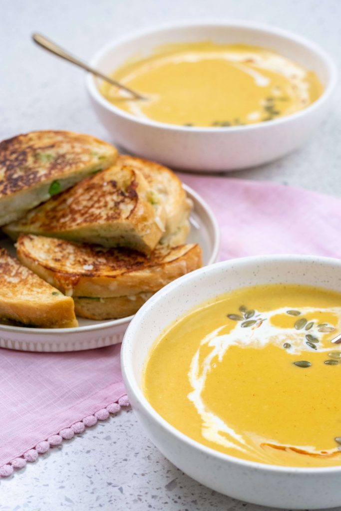 A pile of samosa-style grilled cheese sandwiches made with B.C. cheese and bread on a plate beside two bowls of creamy B.C. pumpkin soup.