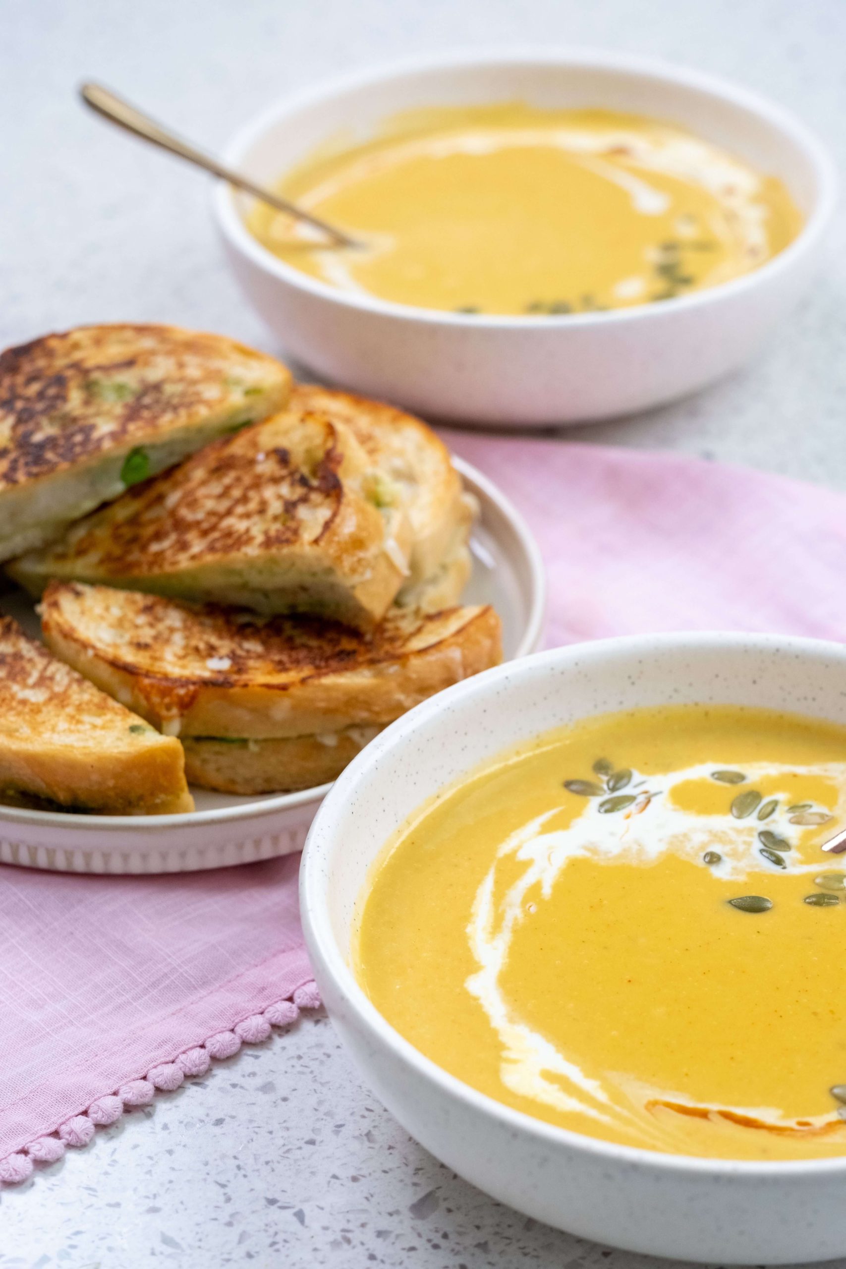 A pile of samosa-style grilled cheese sandwiches made with B.C. cheese and bread on a plate beside two bowls of creamy B.C. pumpkin soup.