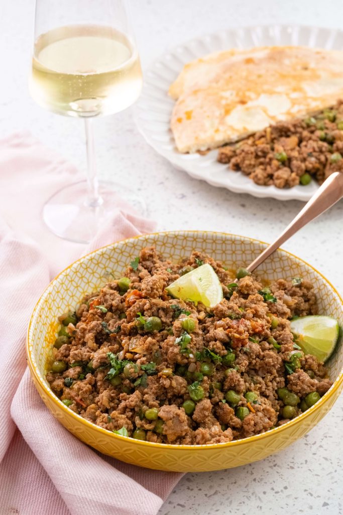A yellow bowl filled with ground B.C. lamb keema garnished with lime wedges, sitting on a soft pink napkin next to a glass of sparkling B.C. wine, and a plate of naan bread in the background. 