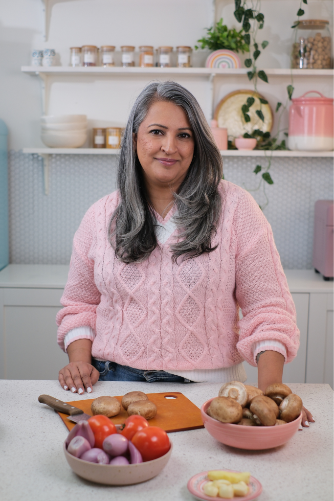Raj Thandhi standing in the kitchen with ingredients, mushrooms, garlic, and tomato. 